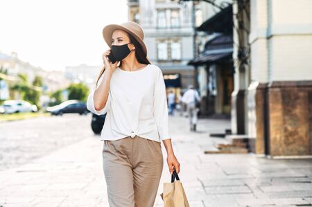 A young woman in a protective mask walks along a city street with a paper shopping bag in her hands and speaks on the phone. City walking and shopping after quarantineの写真素材