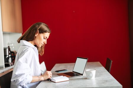 Side view young woman using laptop for studying or works and takes notes while sitting indoor at homeの写真素材