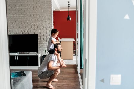 Sport at home with a child. Young dad does squats in cozy living room, his cute toddler son sits on his shoulders. Side viewの写真素材