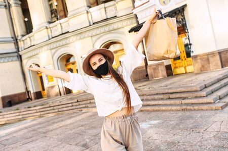 The girl is happy with the end of quarantine, she walks around the city. Young happy woman with a protective mask on her face raised her hands up, a shopping bag in her handの写真素材
