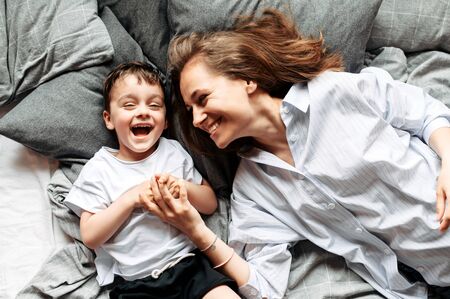 Mom and little cute child have fun at home. Young mom and toddler son lie on the bed and laughing. Top viewの写真素材