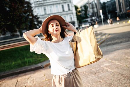 Girl enjoys a walk in the city. Young beautiful girl in a hat and with a paper shopping bag stands in the city center with pleasure closed her eyesの写真素材