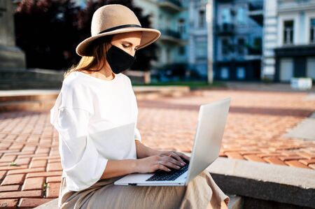 A girl in a protective mask on her face in casual clothes sits on the steps in the city and uses a laptop.の写真素材