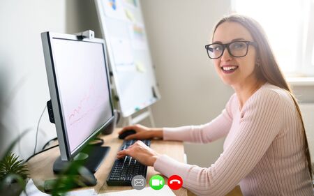Attractive girl in glasses sits at the office desk with PC and looks at camera while talking online via video callの写真素材