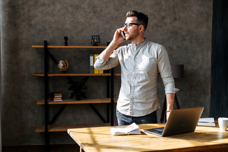 A guy is talking on the phone in the office wearing glasses, he is looking through the window, work place on the background. Young man wears casual clothes in a cosy office roomの写真素材