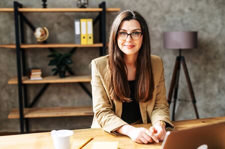 Young brunette successful woman sits at the office desk, she looks into the camera wearing glasses...