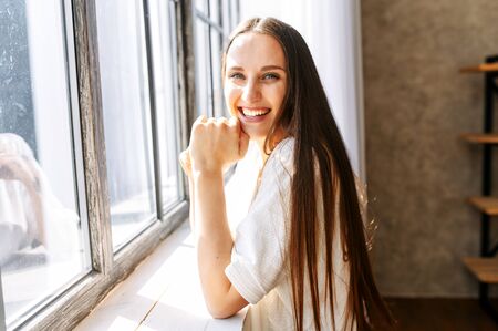 Photo indoor of cheerful young long-haired woman in white t-shirt in relaxed pose looks at camera and laughsの写真素材