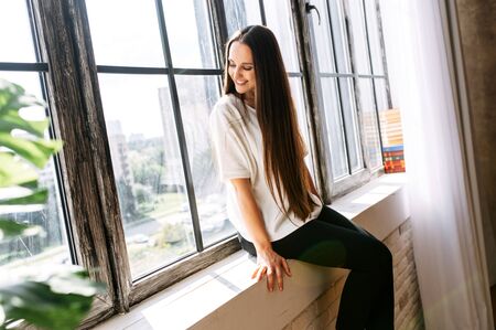 Young woman with long hair in white t-shirt sits relaxed on windowsill. Photo in natural lightの写真素材