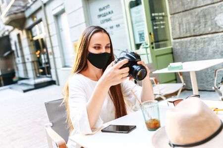 Young woman in medical mask with a camera sits at summer cafe and is watching shots after walking in the city. Tourism after quarantineの写真素材
