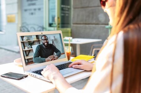 Video call outdoors. A young woman sits in the summer cafe and uses laptop for video connection with a friend, coworker, she talks with an african-american guyの写真素材