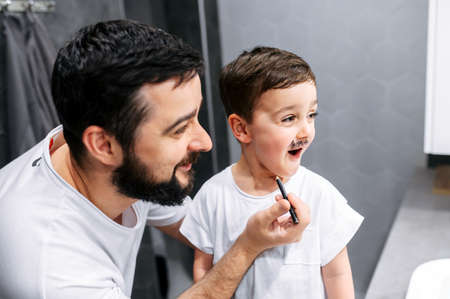 Father and son have a fun in bathroom. Bearded dad is drawing beard on face of little son with a marker and going to shave him, they laughの写真素材