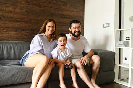 A young family spends home leisure watching TV. Happy mom, dad and little son are sitting behind a sofa in a cozy living room. Photo with grainの写真素材