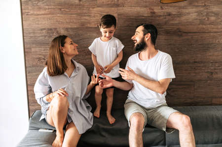 Happy mom and dad are sitting on the couch in the cozy living room and looking with love at their son. Family time. Photo with grainの写真素材