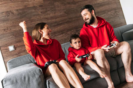 Home entertainment with playing console. Father, mother and little son with a joysticks sits on the cozy couch at home and play on play station. Photo with grainの写真素材