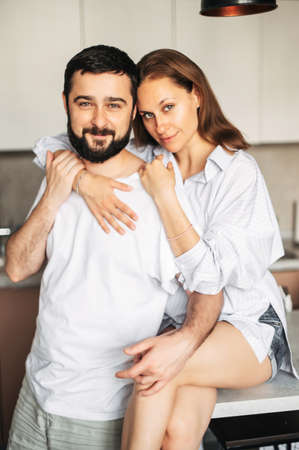 A married couple in love hugging in the kitchen. A beautiful young wife sits on the edge of the table, her husband stands near, they look at camera. Vertical shotの写真素材
