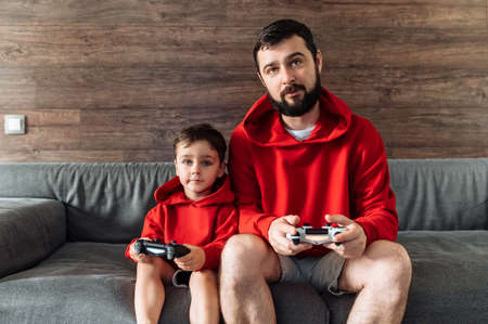 Home leisure with playing console. Father and little son with a joysticks sits on the cozy couch at home and play on play station. Photo with grainの写真素材