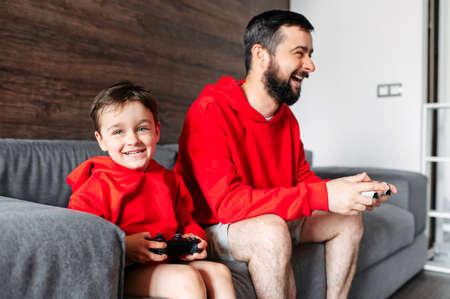 Father and toddler spend home leisure with play station. Dad and boy son in red hoodies sit on the sofa with joysticks in their hands and play on the console with excitement. watching TV. Photo with grainの写真素材