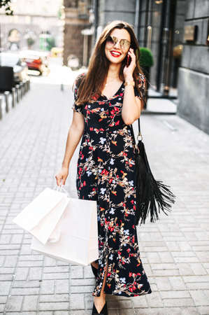 Vertical shot of attractive brunette girl in long summer dress outdoors, girl wearing sunglasses with a shopping bags is walking among stores in the city and talking on the phoneの写真素材