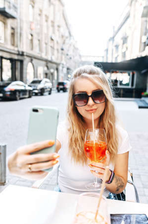 Fancy blonde girl in sunglasses drinks a cocktail and takes a selfie on the trendy smartphone while sitting outdoor in the summer cafe. Vertical shotの写真素材