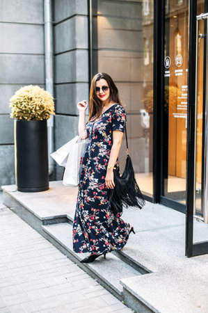 Attractive girl comes out of the store with shopping bags after shopping. Brunette in a long dress comes down on stairs with paper shopping bags in her hand.の写真素材
