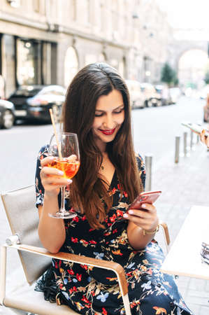 Portrait of beautiful young brunette girl with cocktail in hands, woman sits in the summer cafe and using phone for chattingの写真素材
