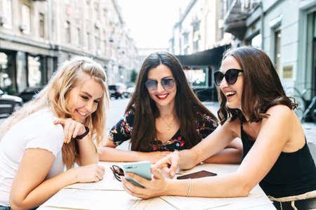 Cheerful ladies in the summer cafe looks on smartphone screen and have lively discussion. Women talk about the news, gossip.の写真素材