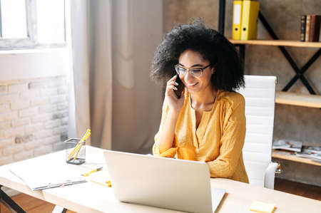 Charming biracial young woman in eyeglasses talks on the smartphone while working with a laptop in modern officeの写真素材