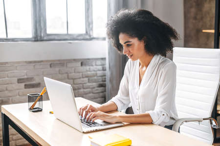 Concentrated biracial girl uses laptop for work in the modern office. Beautiful young woman types on a keyboard and looks at the computer screenの写真素材