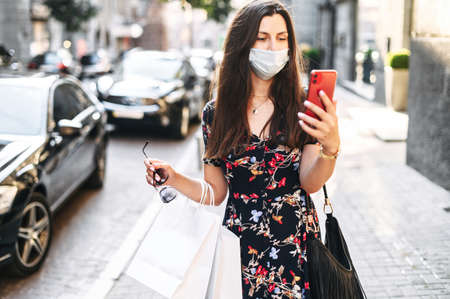 Protect yourself from viruses, allergies, smog. The girl with a protective medical mask on the face goes on the street after shopping with paper packages and looks at smartphone screenの写真素材