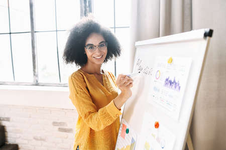 A young mixed-race woman in eyeglasses is writing on flipchart, looks at camera and smiles. Online teacher, tutor. Side viewの写真素材