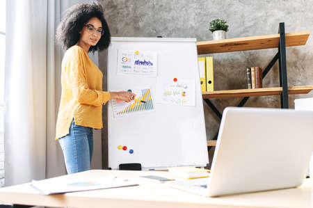 Confident young african-american woman conducts webinar, business training online. Online coach stands near flip chart in front of laptop and explains something to online audienceの写真素材