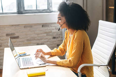 Young attractive girl uses headset for talk online while working in the light modern office. Side view of an ethnic woman with an afro hairstyle in the officeの写真素材