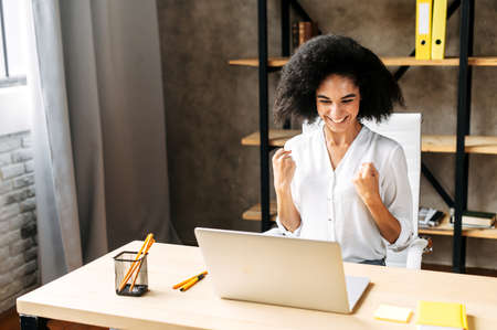 Happy attractive young biracial woman in modern office had seen good news in network, she looks at screen and clenched her fists in triumph. Victory conceptの写真素材