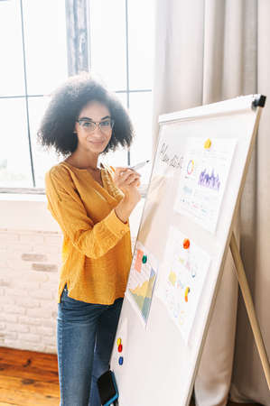 A young mixed-race woman in eyeglasses is writing on flipchart, looks at camera and smiles. Online teacher, tutor. Side viewの写真素材