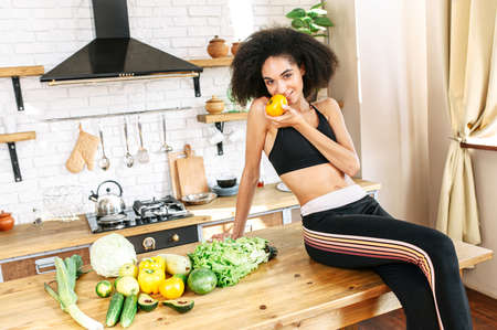 Young attractive african woman is sitting on the countertop in the kitchen with a fresh yellow tomato in her hands, looking at camera and smiling. Fresh vegetables near on the table. Healthy and wholesome foodの写真素材
