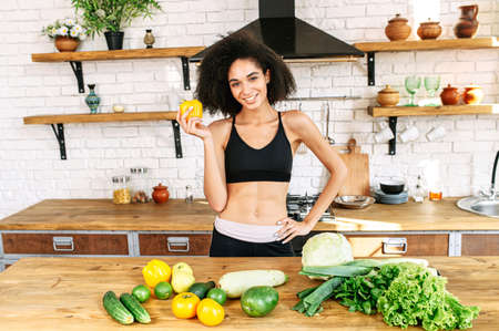 Weight loss diet. Attractive african-american woman with a athletic fit body stands in the kitchen near table full of vegetables on it, holds yellow pepper in hands and looks at camera with a smileの写真素材