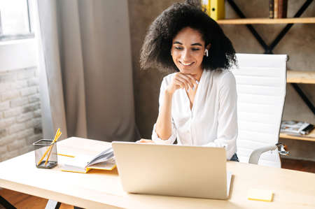 Pretty biracial girl is office employee. Attractive girl in white shirt uses earphones for talking online via video call on the laptopの写真素材
