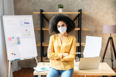 Mixed-race woman with a medical mask on a face stands in office and looks at camera with arms crossed. Viral protection indoorの写真素材