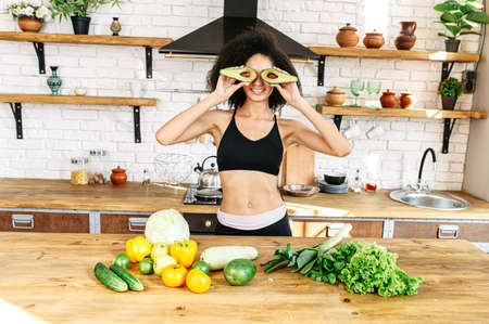 African-american woman with an afro hairstyle covered her eyes with halves of avocado, she stands in the kitchen near table with vegetables on it. Funny picture with healthy food. Keto diet conceptの写真素材