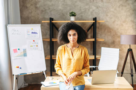 Attractive biracial woman with a medical mask in hands stands in office and looks at camera. Portrait of charming mixed-race girl with an afro hairstyle indoor.の写真素材