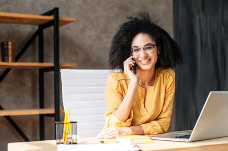 A friendly biracial young woman sits at the office desk and talks on the phone, she looks at camera with a positive smile. Friendly office atmosphereの写真素材