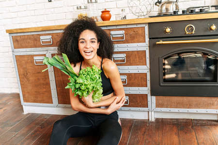 Cheerful biracial girl with an armful of greens in her hands sits on the floor in the kitchen. A girl with an Afro hairstyle in sportswear holds salad, leek, basil and other herbs, looks at camera and laughsの写真素材