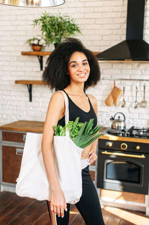 Healthy lifestyle concept. Young sporty african-american woman in the kitchen with a shopping bag full of fresh vegetablesの写真素材