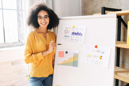 An african-american female woman conducts business training. Young woman uses flip chart for explaining. A smiling girl points on whiteboard and looks at camera with a smile. Front viewの写真素材