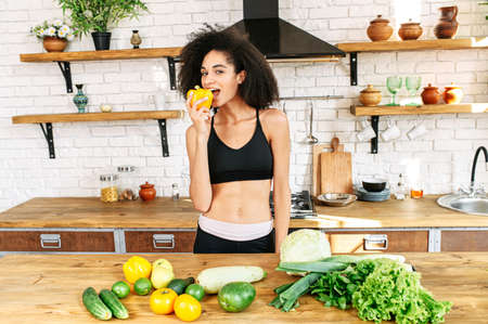 Eat healthy foods. Athletic african american girl in sportswear in the kitchen bites fresh pepper, vegetables and greens on the tableの写真素材