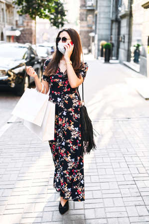 Shopping with protective measures after quarantine is ended. Woman walks on the city street with a medical mask on the face and talks on the phone, paper shopping bags in her handsの写真素材