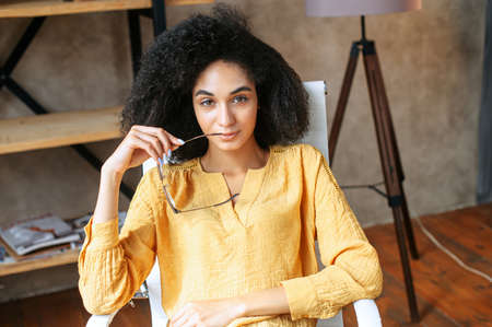 Smart and attractive young mixed-race woman put off eyeglasses, sits in office chair and looks at camera confidentlyの写真素材
