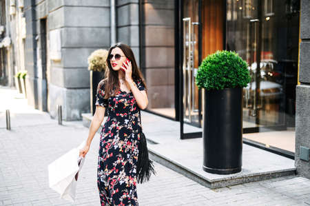 Pretty young woman in long summer dress walks outdoors and talks on the phone after shopping day, paper bags with purchases in her handの写真素材