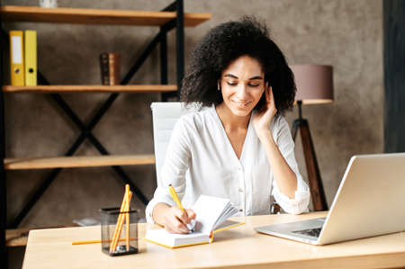 Positive girl is office employee. Attractive girl in white shirt in office uses smartphone for connection with customers, she takes notes while speakingの写真素材