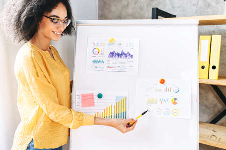 An african american female teacher, tutor, mentor. A young woman in eyeglasses with an afro hair points on flip chartの写真素材